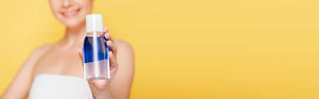 selective focus of smiling beautiful woman holding micellar water in bottle isolated on yellow, panoramic shotの写真素材