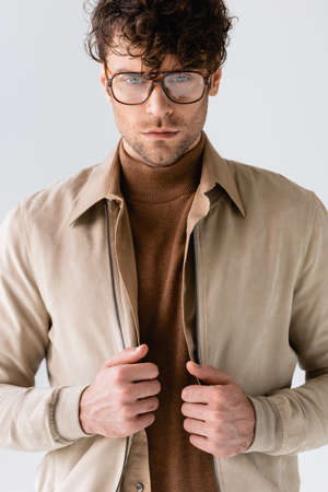 serious, fashionable man in eyeglasses touching jacket isolated on gray