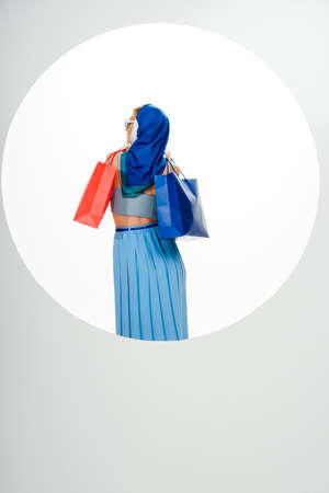 Back view of stylish woman in headscarf holding red and blue shopping bags near circle on white backgroundの写真素材