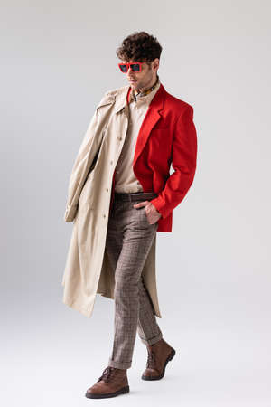 full length view of trendy man with trench coat over shoulder posing on gray with hand in pocketの写真素材