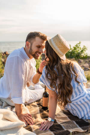happy man touching face of cheerful woman in straw hat near bottle with wineの写真素材
