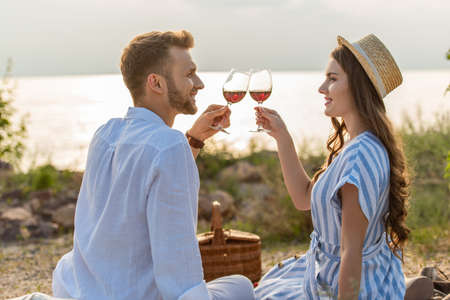 side view of cheerful couple clinking glasses with red wine while sitting near lakeの写真素材
