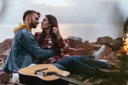selective focus of bearded man hugging girlfriend near acoustic guitar and bonfire