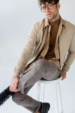 stylish, confident man looking at camera while sitting on stool and touching leg isolated on grayの写真素材