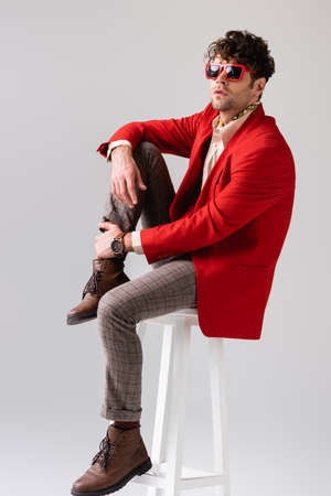 fashionable man in red blazer and sunglasses touching leg while sitting on stool isolated on grayの写真素材