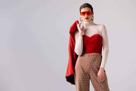 fashionable girl in sunglasses holding red blazer while looking at camera isolated on gray