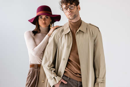 full length view of stylish woman in hat touching trendy man posing with hand in pocket isolated on...