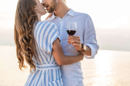 cropped view of bearded man kissing girlfriend and holding glass with red wine near lakeの写真素材