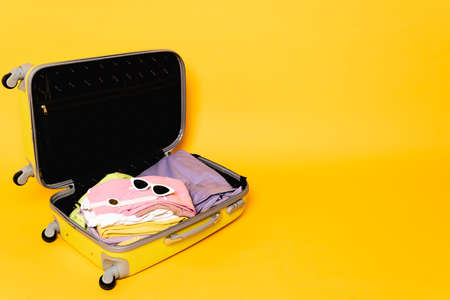 open travel bag with summer clothes and accessories on yellow background