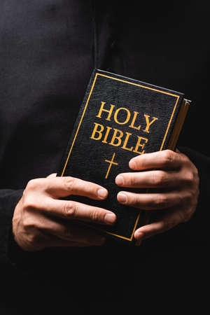 partial view of priest holding holy bible in hands isolated on blackの写真素材