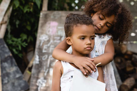poor african american child holding pencil and hugging sad brother in torn clothesの写真素材