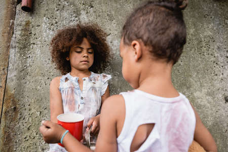Selective focus of destitute afican american kid holding metal plate near brother and concrete wall on urban streetの写真素材