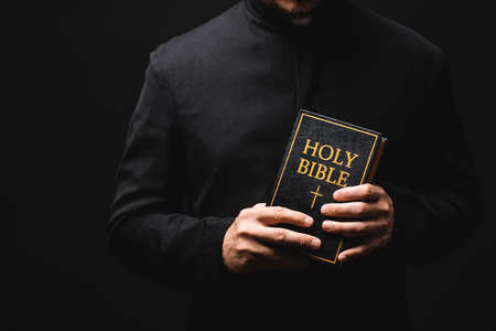 cropped view of priest holding holy bible in hands isolated on blackの写真素材
