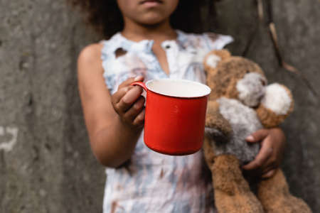 Cropped view of poor african american kid holding metal cup and teddy bear while begging alms on urban streetの写真素材