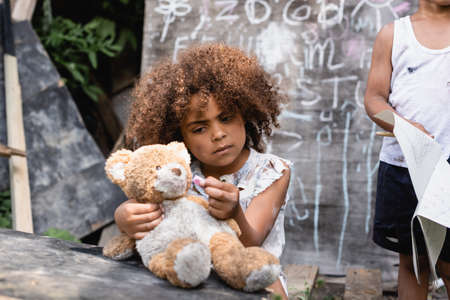 selective focus of poor african american kid looking at dirty teddy bear near child and chalkboardの写真素材