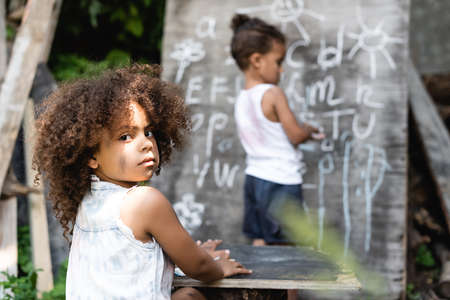 selective focus of curly african american kid sitting near table, boy and chalkboardの写真素材