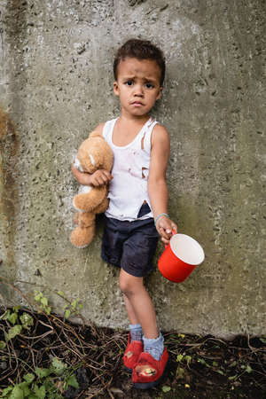 Dissatisfied african american child in dirty clothes begging alms near concrete wall in slumの写真素材