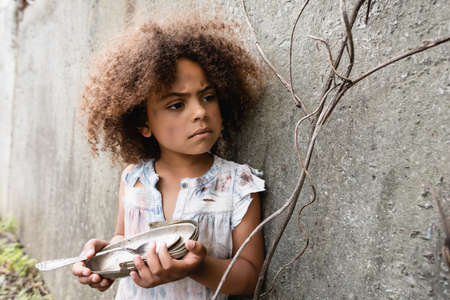 Selective focus of poor african american kid holding dirty metal plate and spoon near concrete wall on urban streetの写真素材