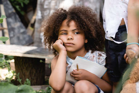 poor african american kid holding notebook near child standing with dirty soft toy