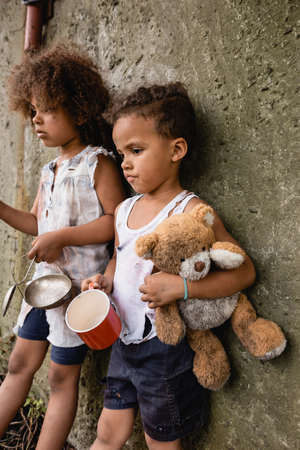 Selective focus of sad african american boy holding dirty teddy bear while begging alms near sister and concrete wall in slumの写真素材