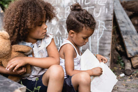 selective focus of poor african american kid looking at brother holding notebook and pencilの写真素材