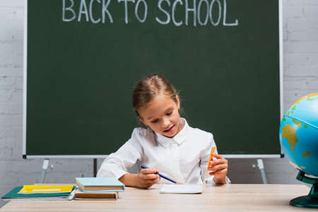 attentive schoolgirl looking in copy book while sitting at desk near textbooks and chalkboard with...