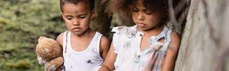 Panoramic crop of sad homeless afican american kids with dirty teddy bear standing near concrete wall in slumの写真素材
