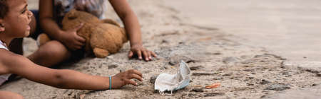 panoramic crop of poor african american boy crying while reaching dirty medical mask near sister on groundの写真素材