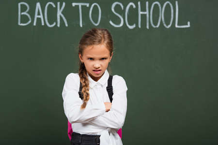 angry schoolgirl standing with crossed arms near chalkboard with back to school letteringの写真素材