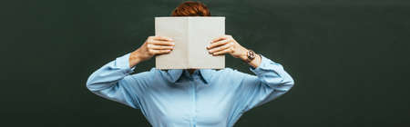 panoramic crop of teacher obscuring face with open book while standing near chalkboardの写真素材