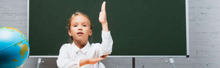 panoramic crop of adorable schoolgirl sitting with raised hand near globe and chalkboard
