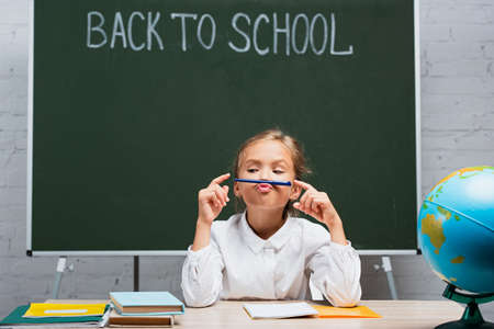 cute schoolgirl holding pen near lips while sitting at desk near globe and chalkboard with back to school letteringの写真素材