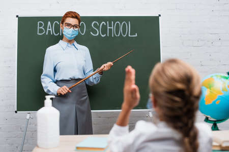 back view of schoolgirl with raised hand and teacher with pointing stick near chalkboard with back to school letteringの写真素材