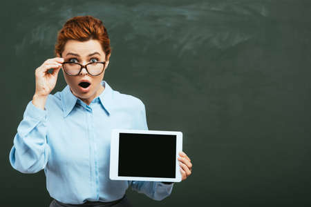 shocked teacher touching glasses while holding digital tablet with blank screen near chalkboard
