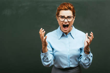 angry teacher in eyeglasses gesturing and screaming near chalkboardの写真素材