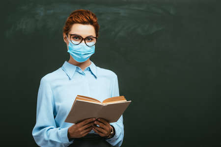 young teacher in eyeglasses holding open book and looking at camera while standing near chalkboard