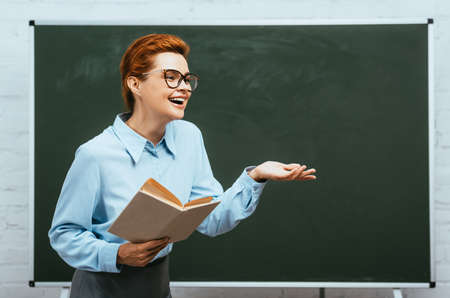 laughing teacher in eyeglasses holding book while standing near chalkboard with open armの素材 [FY310152617294]