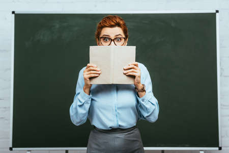 scared teacher in eyeglasses covering face with open book while standing near chalkboardの写真素材