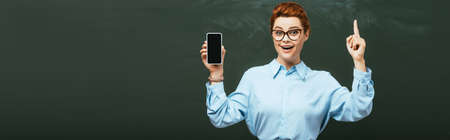 horizontal concept of smiling teacher showing idea gesture while holding smartphone with blank screen near chalkboardの写真素材