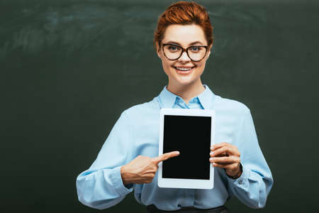happy teacher pointing with finger at digital tablet with blank screen near chalkboardの写真素材