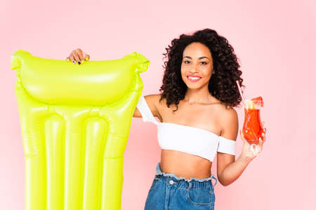 happy african american girl standing near inflatable mattress and holding cocktail isolated on pinkの写真素材