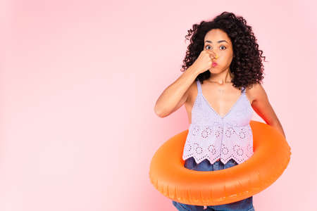 african american woman standing with swim ring and touching nose isolated on pink