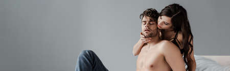 Panoramic shot of woman touching chin of shirtless boyfriend on bed on gray background