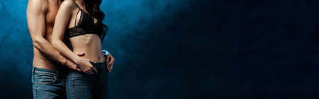 Panoramic crop of muscular man embracing woman in jeans and bra on black background with smoke