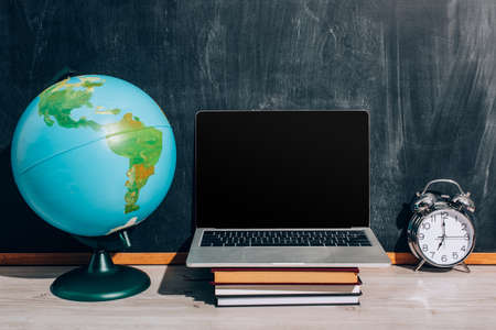 laptop with blank screen on stacked books, globe and alarm clock near black chalkboardの写真素材