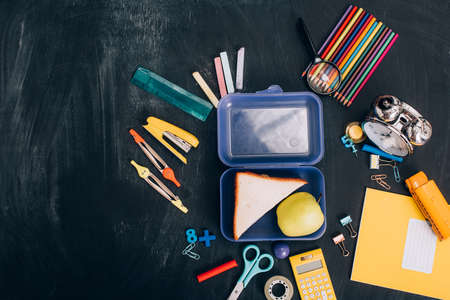 top view of lunch box with whole apple and tasty sandwiches near school stationery on black...