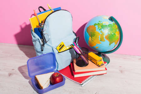 blue backpack with school supplies near globe, lunch box, books, pen holder and school bus model on pinkの写真素材