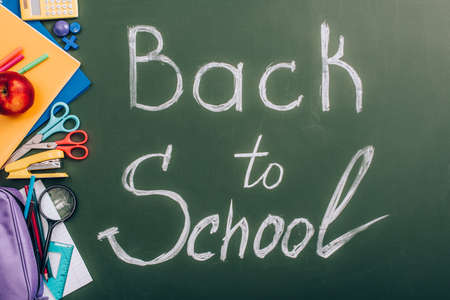 top view of back to school inscription near school supplies and fresh apple on green chalkboard
