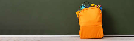 horizontal image of yellow backpack full of school stationery on desk near green chalkboard