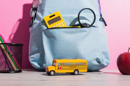 blue backpack with school supplies near school bus model, ripe apple and pen holder with felt pens on pinkの写真素材